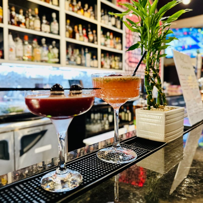 Two cocktails, one red and one orange, on a bar with a liquor shelf backdrop at Bazil Indian Cuisine.