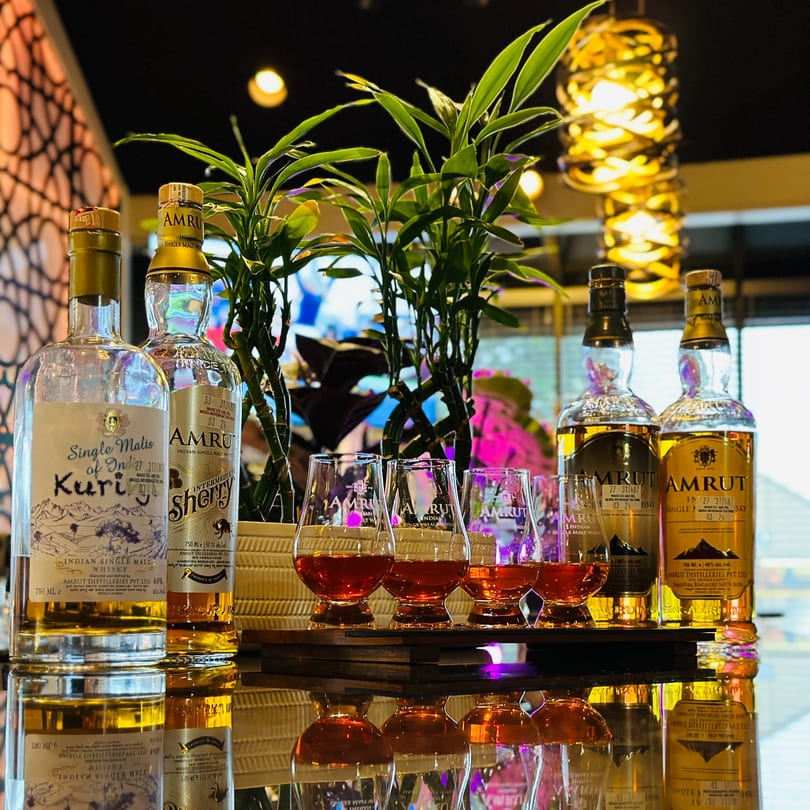 Six bottles of Amrut Indian single malt whisky and four glasses with amber liquid on a reflective surface.