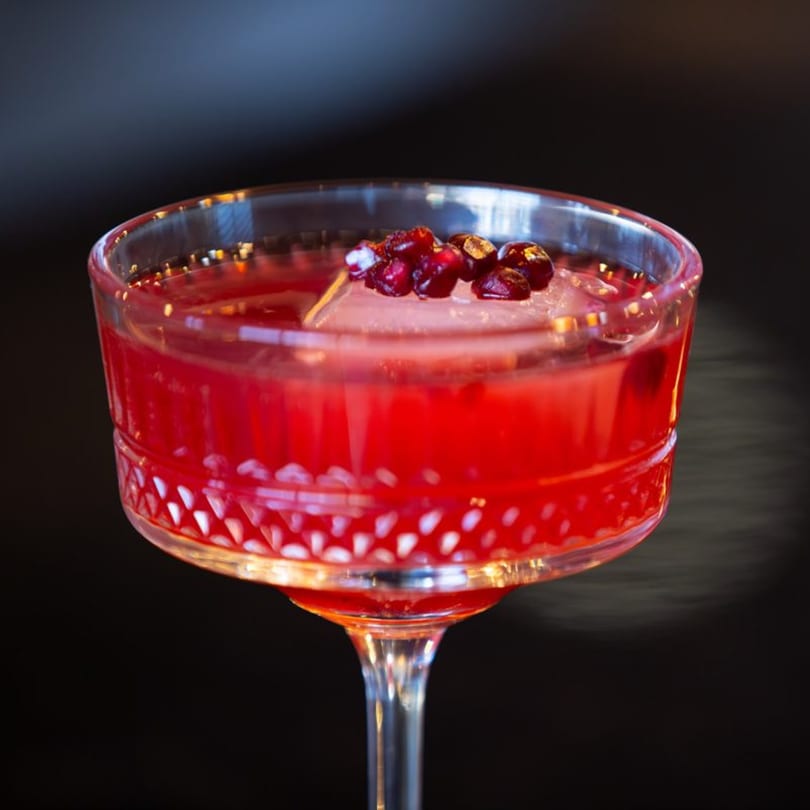 A vibrant red cocktail in a stemmed glass, garnished with pomegranate seeds and an ice cube.