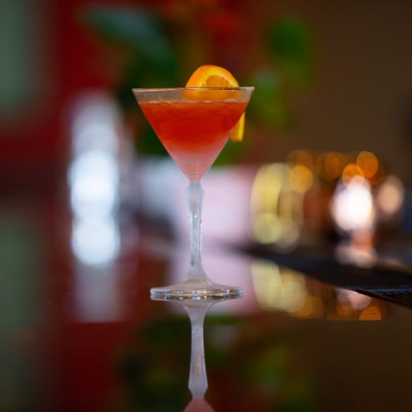 A vibrant red cocktail in a martini glass, garnished with an orange slice, on a reflective surface.