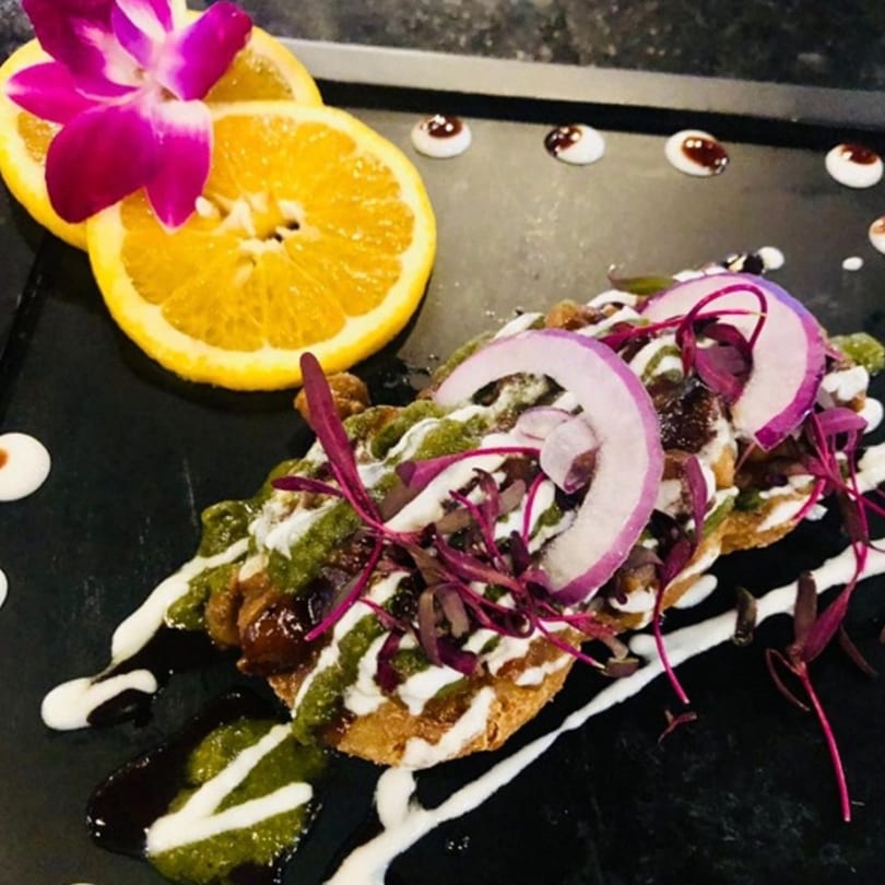 Indian chaat with red onions, green sauce, and white drizzle, garnished with orange slices and an orchid.