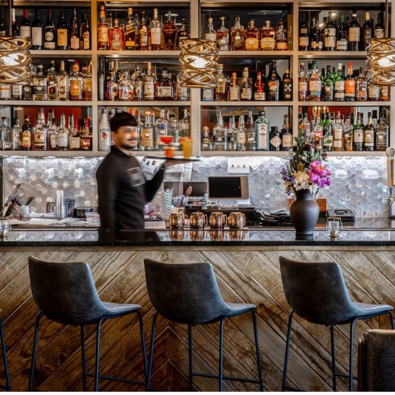 A bartender at Bazil Indian Cuisine carries drinks behind a well-stocked bar with bar stools.
