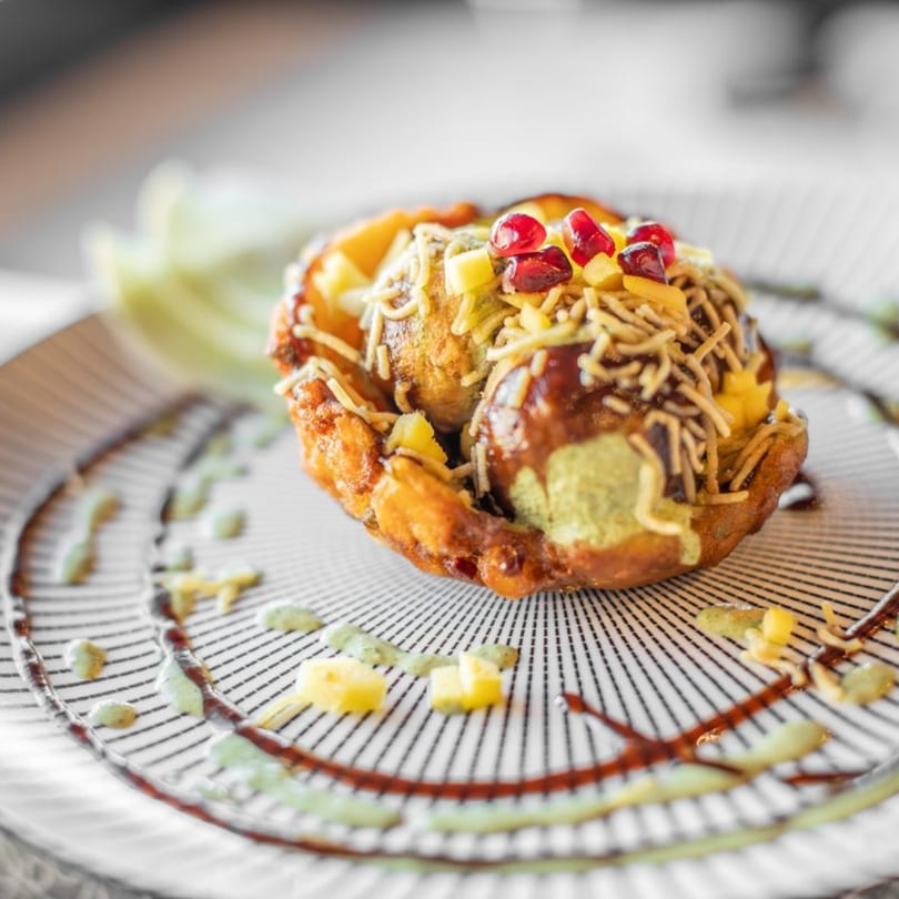 Close-up of a golden-fried Indian dish topped with pomegranate, cheese, and sauces on a striped plate.