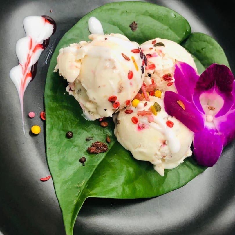 Three scoops of Indian ice cream, garnished with colorful sprinkles, a purple orchid, and a green leaf.
